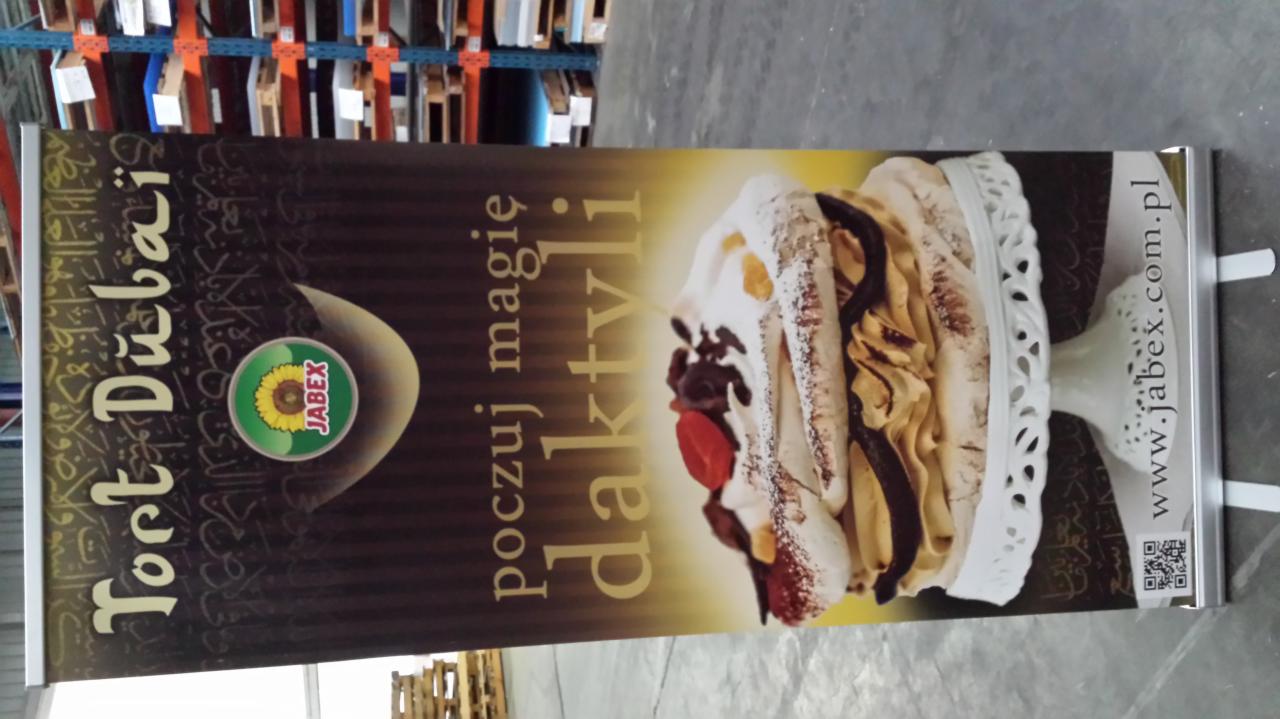 Roll-up reklamowy z grafiką tortu daktylowego i logo Jabex, umieszczony w magazynie. Widoczne regały w tle. Całość obrócona o 90 stopni w lewo.