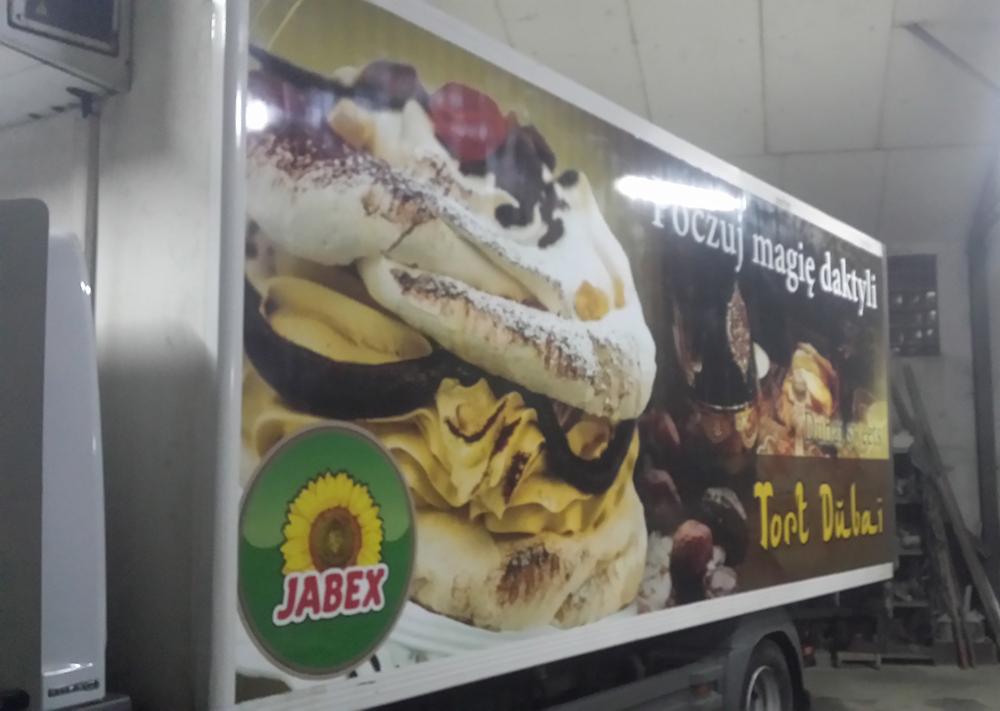 Reklama nadrukowana na boku ciężarówki z tortem 'Tort Dubai' i logo Jabex, prezentująca słodkie produkty i hasło 'Poczuj magię daktyli'.