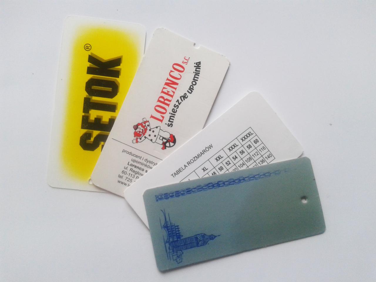 Stos etykiet odzieżowych różnych marek, w tym z tabelą rozmiarów i logo firmy odzieżowej, na białym tle. Widoczne nazwy firm i adresy.