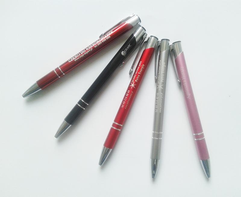 Zestaw pięciu długopisów reklamowych w różnych kolorach (czerwony, czarny, srebrny, różowy) z nadrukowanymi logotypami i danymi kontaktowymi firm na białym tle.