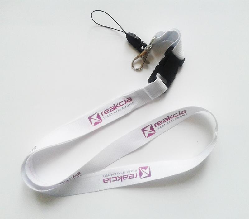 Biała smycz reklamowa z fioletowym nadrukiem logo firmy 'Reakcja Flagi Reklamowe', z czarnym zapięciem i zawieszką na telefon, leżąca na białym tle.