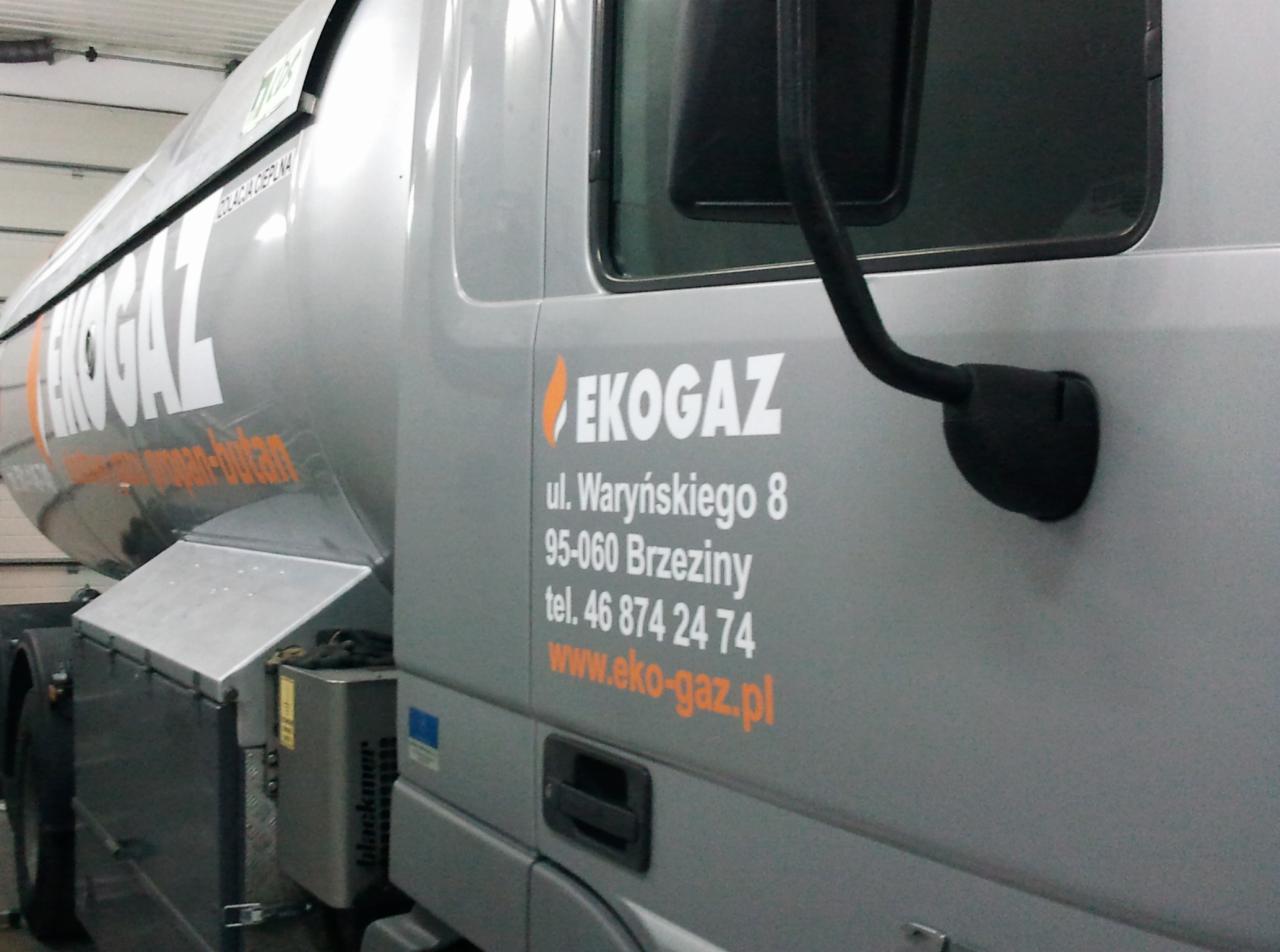 Cysterna EkoGaz z nadrukowanym logo i danymi kontaktowymi firmy na szarym tle. Widoczny fragment opony i lusterko boczne. Całość kadru wypełnia bok pojazdu.