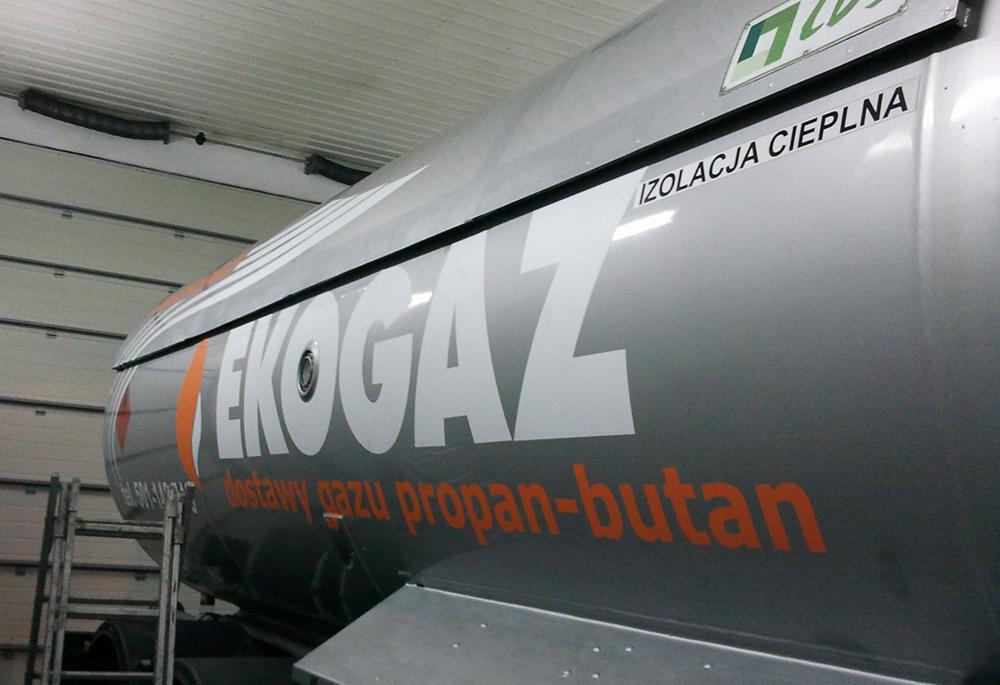 Cysterna z napisem 'EKOGAZ' i informacją 'dostawy gazu propan-butan' w pomarańczowym kolorze, widoczna drabina i fragment hali.