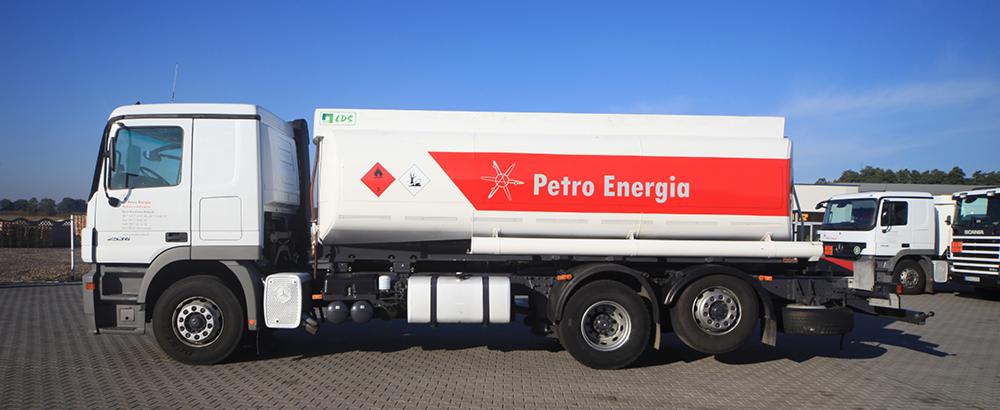 Biała cysterna z czerwonym logo Petro Energia na tle błękitnego nieba. Widoczne oznaczenia substancji niebezpiecznych. Na pierwszym planie kostka brukowa.