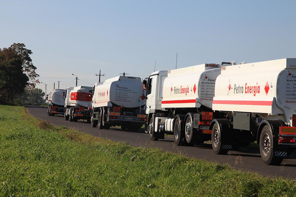 Kolumna białych cystern z czerwonym logo 'Petro Energia' na drodze, widok z boku.  Cysterny jadą w jednym kierunku, po prawej stronie drogi zielona trawa.