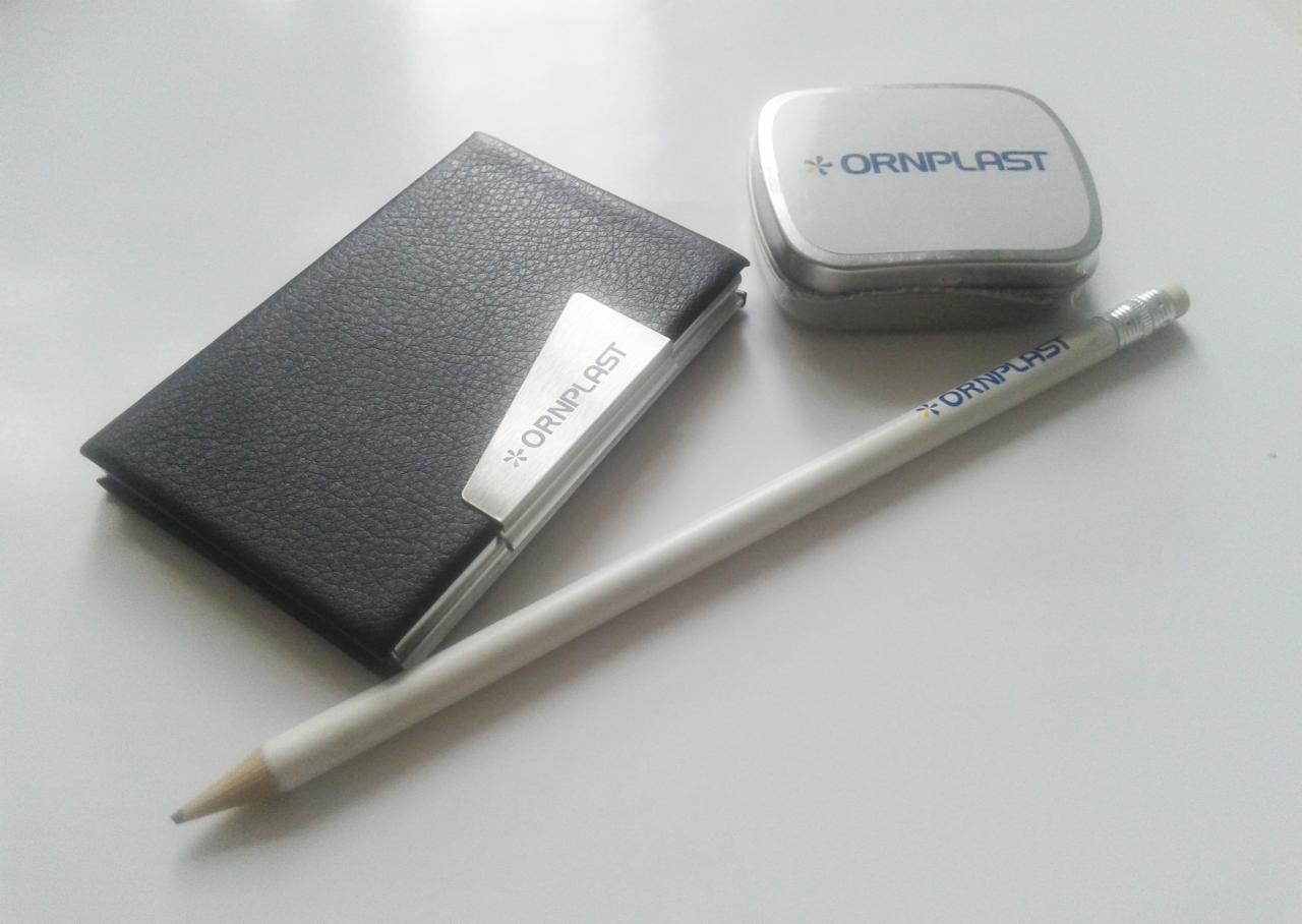 Czarny wizytownik skórzany z metalową klamrą, ołówek i metalowe pudełko z nadrukiem logo firmy ORNPLAST na białym tle.