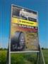 Billboard reklamowy z grafiką opon na tle błękitnego nieba i zielonej trawy. Plakat stoi na metalowym stelażu. Ujęcie z dołu, podkreślające wielkość reklamy.