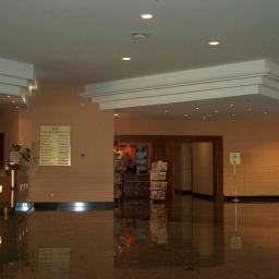 WARM-DOM SP. Z O.O. - Przestronne lobby hotelowe z marmurową posadzką, recepcją po lewej, tablicą informacyjną i windą po prawej stronie, oświetlone punktowymi lampami sufitowymi.