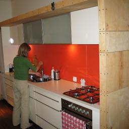 WARM-DOM SP. Z O.O. - Kobieta myje naczynia w zlewie w kuchni z pomarańczowym panelem ściennym i surową drewnianą konstrukcją szafek.