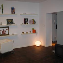 WARM-DOM SP. Z O.O. - Jasne wnętrze z ciemną podłogą, białymi ścianami i półkami z dekoracjami, oświetlone kulistą lampą podłogową, widoczny fotel i stary telewizor.