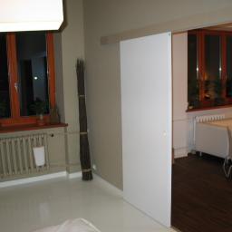 WARM-DOM SP. Z O.O. - Nowoczesne wnętrze z przesuwnymi, białymi drzwiami, widoczne okna z drewnianą ramą, grzejnik ścienny i dekoracyjny snopek gałęzi przy ścianie.