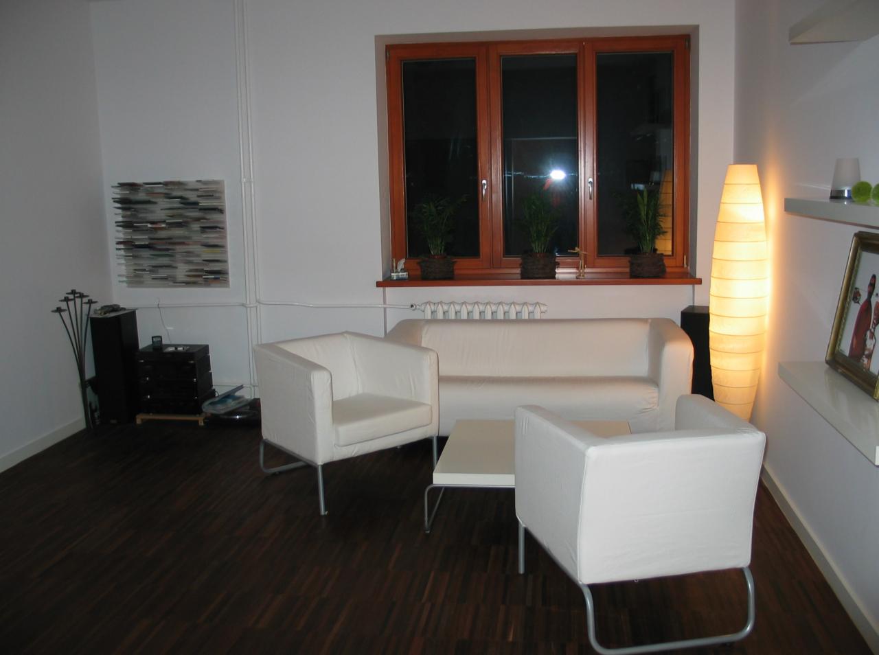 Jasne wnętrze pokoju z białymi meblami: sofą, dwoma fotelami i stolikiem kawowym, na ciemnej drewnianej podłodze, z trzema roślinami doniczkowymi na parapecie okna