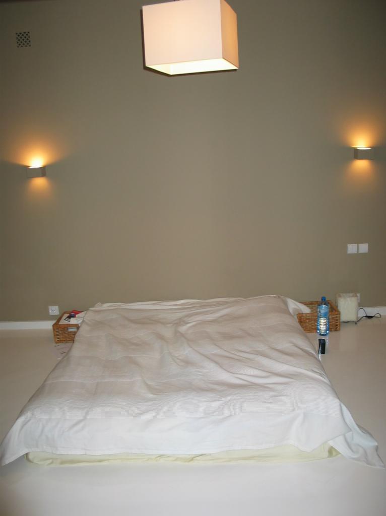 Minimalistyczna sypialnia w Warszawie z łóżkiem na podłodze, oświetleniem ściennym i wiszącą lampą w kształcie sześcianu, ukazująca efekt świeżo pomalowanej ściany w odcieniach beżu.