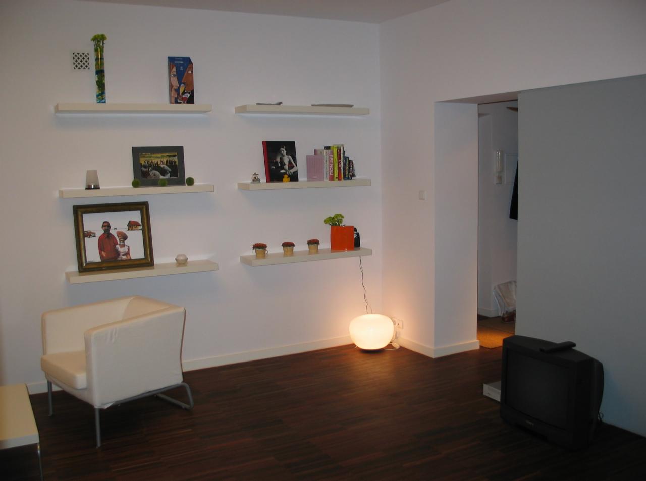 Jasne wnętrze z ciemną podłogą, białymi ścianami i półkami z dekoracjami, oświetlone kulistą lampą podłogową, widoczny fotel i stary telewizor.