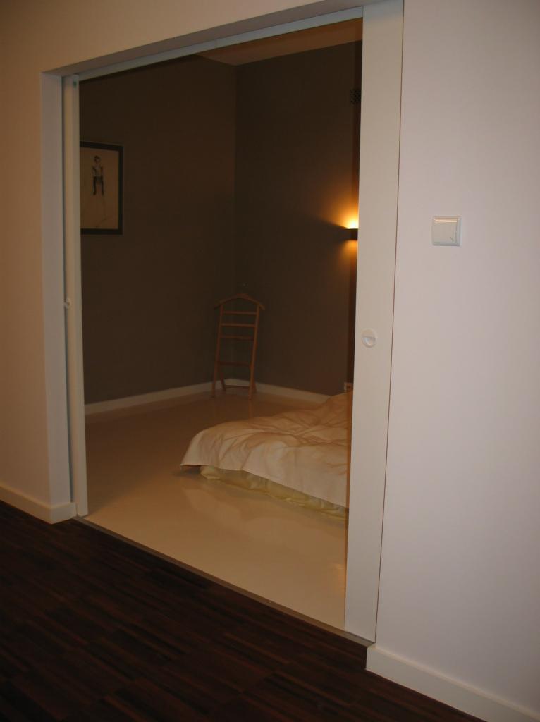 Widok na sypialnię z przesuwnymi drzwiami, beżowa podłoga, brązowe ściany, drewniany wieszak na ubrania, lampka ścienna, obraz na ścianie.