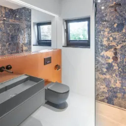 Nowoczesna łazienka z niebiesko-złotymi płytkami imitującymi marmur, szara umywalka nablatowa z szafką, wisząca toaleta w kolorze szarym, miedziana armatura i dwa okna.