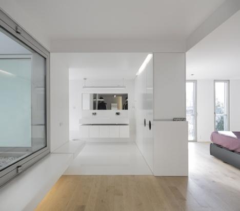 Minimalistyczne, białe wnętrze mieszkania z widokiem na łazienkę i sypialnię, podłoga z jasnego drewna, duże okno po lewej stronie.