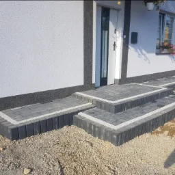 Schody wejściowe do domu wyłożone szarą kostką brukową z białymi fugami, otoczone żwirem, widoczny cień osoby fotografującej.