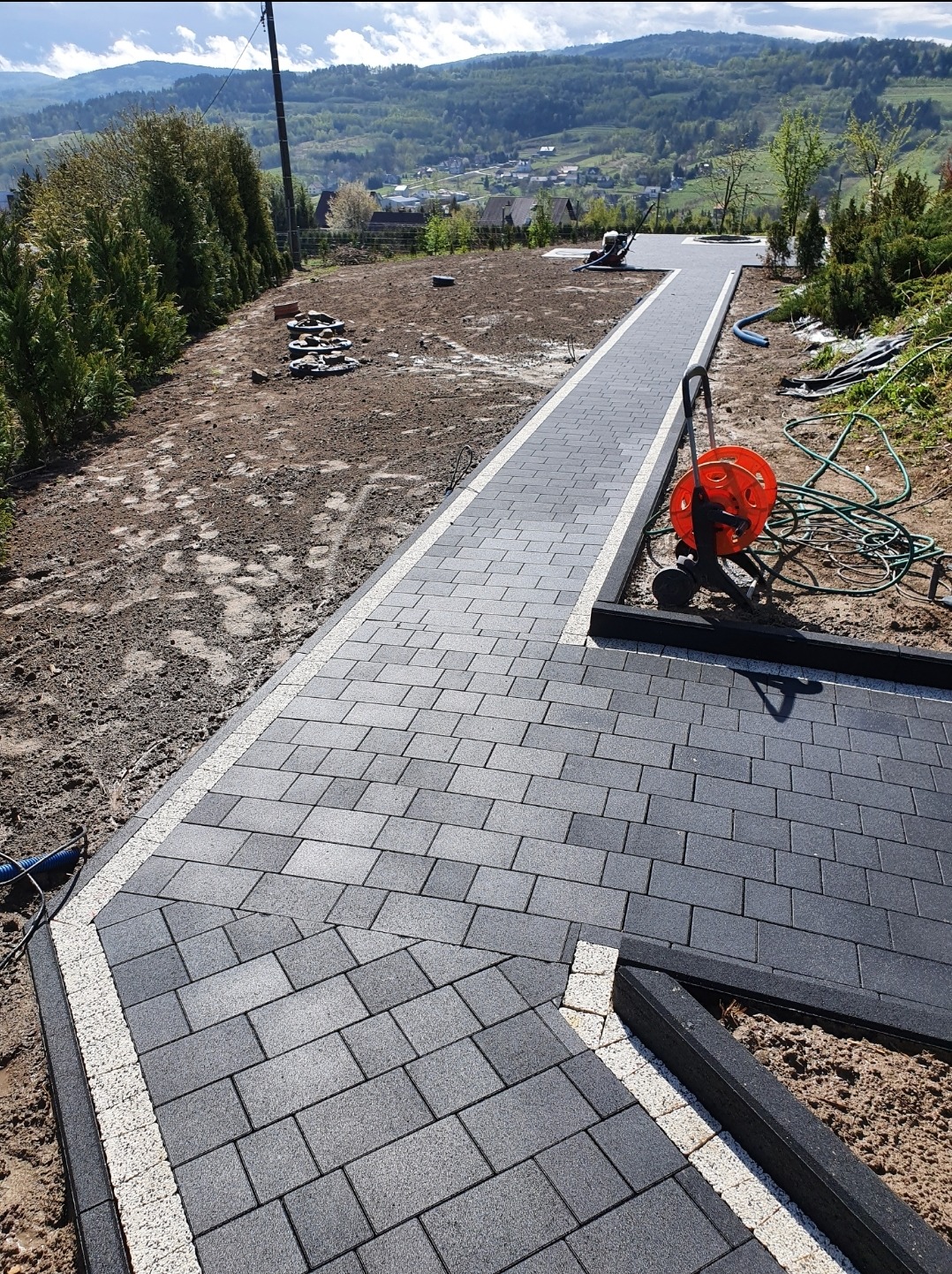 Świeżo ułożona, szara kostka brukowa z białymi obrzeżami na posesji z widokiem na górzysty krajobraz, pomarańczowy zwijak do węża ogrodowego i przygotowany teren pod dalsze prace.