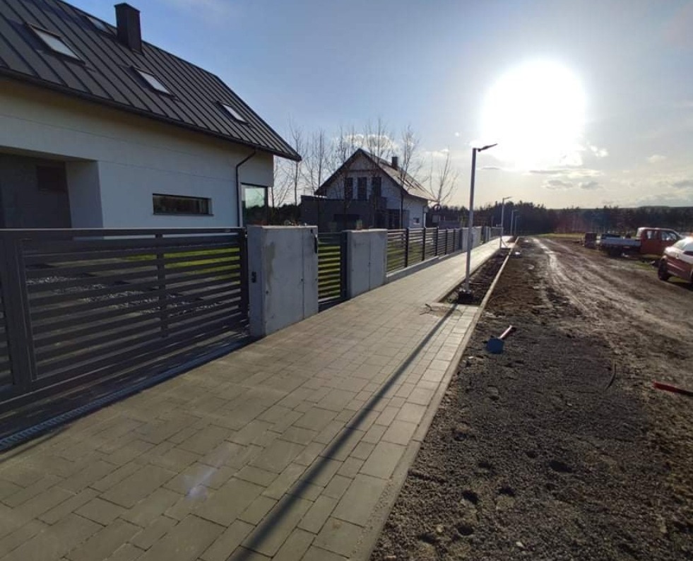 Nowoczesna posesja z szarą kostką brukową prowadzącą do przesuwnej bramy i ogrodzenia z poziomych, metalowych paneli, oświetlona lampami ulicznymi, widok na nieutwardzoną drogę i sąsiednie domy.