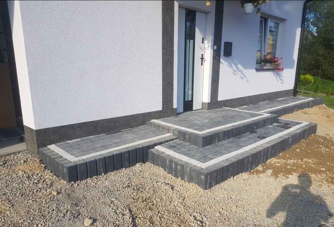 Schody wejściowe do domu wyłożone szarą kostką brukową z białymi fugami, otoczone żwirem, widoczny cień osoby fotografującej.