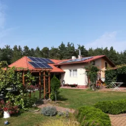 Dom jednorodzinny z panelami słonecznymi na dachu, widoczne również kolektory słoneczne, pergola z roślinnością w ogrodzie, trawnik i drzewa w tle.