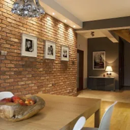Stylowe wnętrze z ceglaną ścianą, ozdobioną trzema obrazami w białych ramkach. Drewniany stół z owocami na pierwszym planie, w tle widoczne schody i regał.