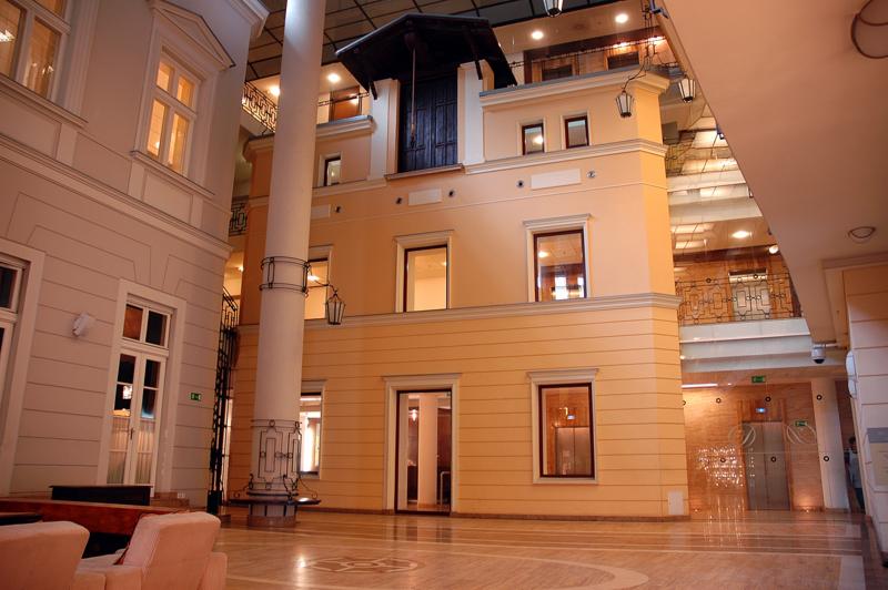 Wnętrze budynku z kolumną i balkonem, widoczne elementy architektoniczne, marmurowa podłoga z wzorem, ciepłe oświetlenie i szklane windy w tle.