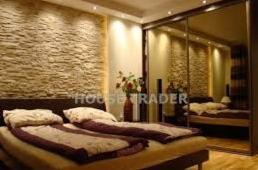 Stylowa sypialnia z kamienną ścianą akcentującą, podświetlona punktowo. Duże lustro w szafie przesuwnych drzwi. Dwuosobowe łóżko z dekoracyjną pościelą.