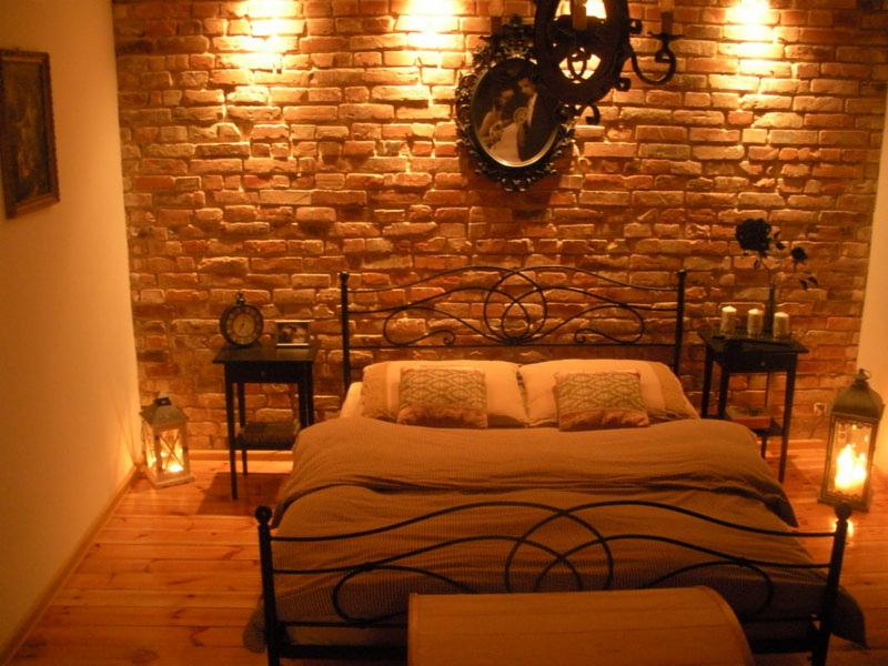 Rustykalna sypialnia z łóżkiem z kutego żelaza na tle ściany z czerwonej cegły, oświetlonej lampami. Klimatyczne lampiony na podłodze i stolikach nocnych.