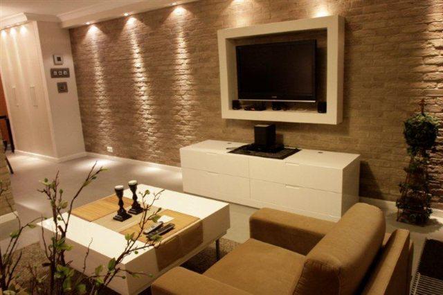 Elegancki salon z telewizorem w białej ramie, kamienna ściana z oświetleniem punktowym, biała komoda i stolik kawowy. Nowoczesne wnętrze z elementami dekoracyjnymi.