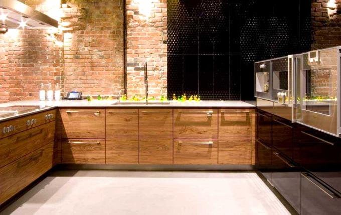 Nowoczesna kuchnia z drewnianymi szafkami, ceglaną ścianą i czarną mozaiką. Widoczne chromowane detale i oświetlenie, tworzące loftowy klimat.