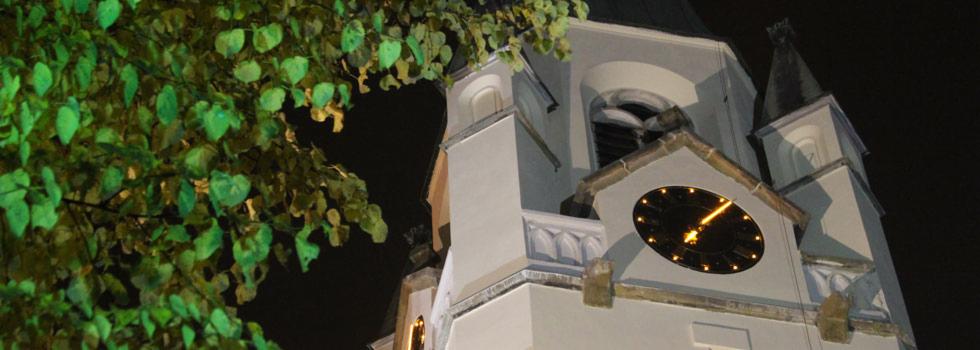 Oświetlona nocą wieża kościoła z zegarem, częściowo zasłonięta przez gałęzie drzewa. Widoczne detale architektoniczne i blask zegara na tle ciemnego nieba.