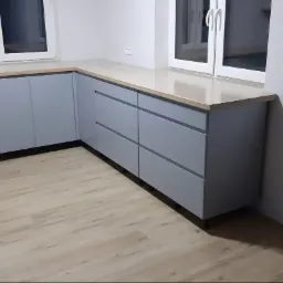 Szare szafki kuchenne z drewnianym blatem pod oknem, widok na fragment podłogi z paneli imitujących drewno.