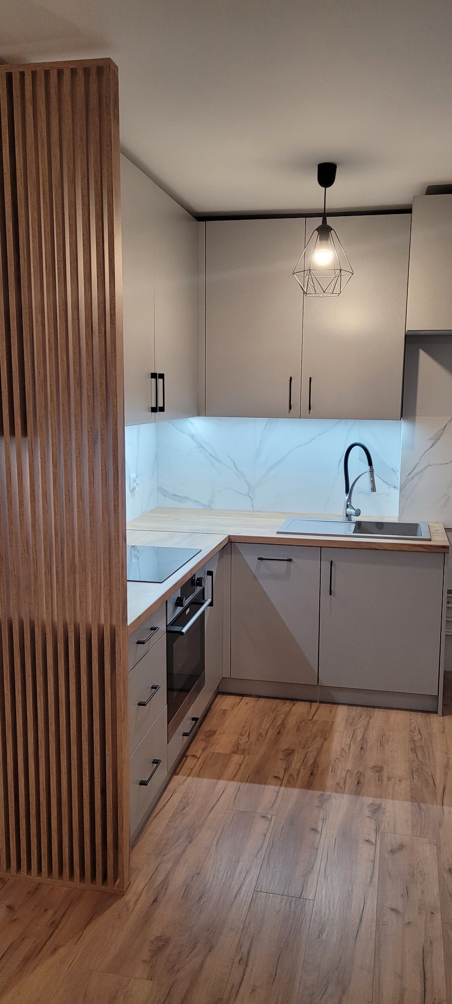 Nowoczesna kuchnia z szarymi szafkami, drewnianym blatem i marmurowym backsplash'em, oświetlona geometryczną lampą wiszącą, z drewnianą ażurową ścianką działową.