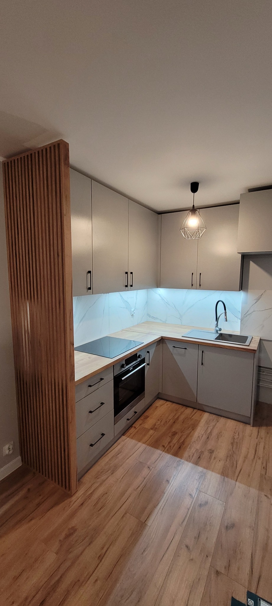 Szara kuchnia w kształcie litery L z drewnianym blatem, marmurowym backsplashiem i drewnianą ażurową ścianką działową.