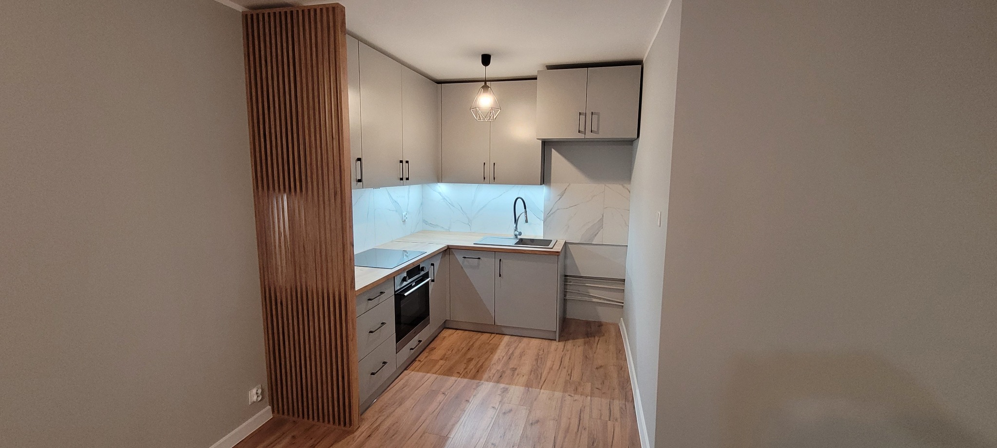 Nowoczesna, jasna kuchnia w kształcie litery U z drewnianym panelem dekoracyjnym, szarymi szafkami i blatem w kolorze drewna. Marmurowe płytki nad blatem, wisząca lampa z metalową konstrukcją.