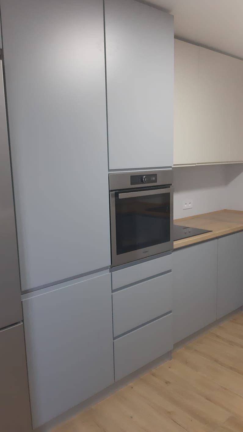 Szare, matowe fronty zabudowy kuchennej z piekarnikiem w zabudowie i drewnianym blatem, widok na fragment podłogi z paneli.