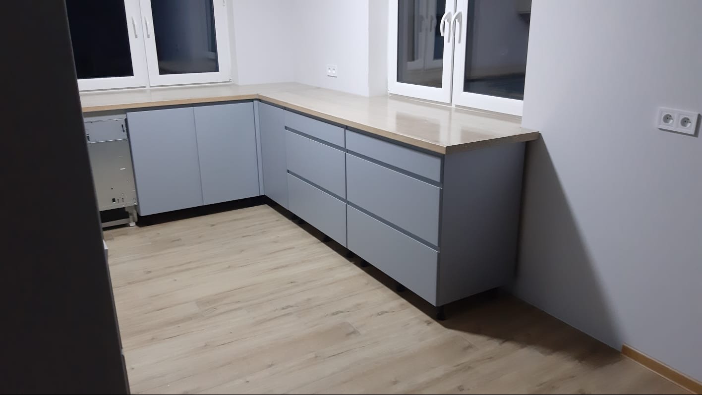 Szare szafki kuchenne z drewnianym blatem pod oknem, widok na fragment podłogi z paneli imitujących drewno.