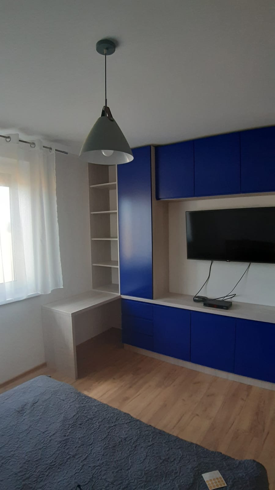 Pokój z zabudową meblową w kolorze kobaltowym, obejmującą szafki, półki i biurko, oświetlony wiszącą lampą z szarym kloszem.