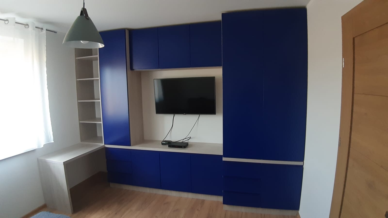 Niebieska zabudowa meblowa z szafkami, półkami i biurkiem, z telewizorem na ścianie, oświetlona wiszącą lampą w pokoju z drewnianą podłogą.
