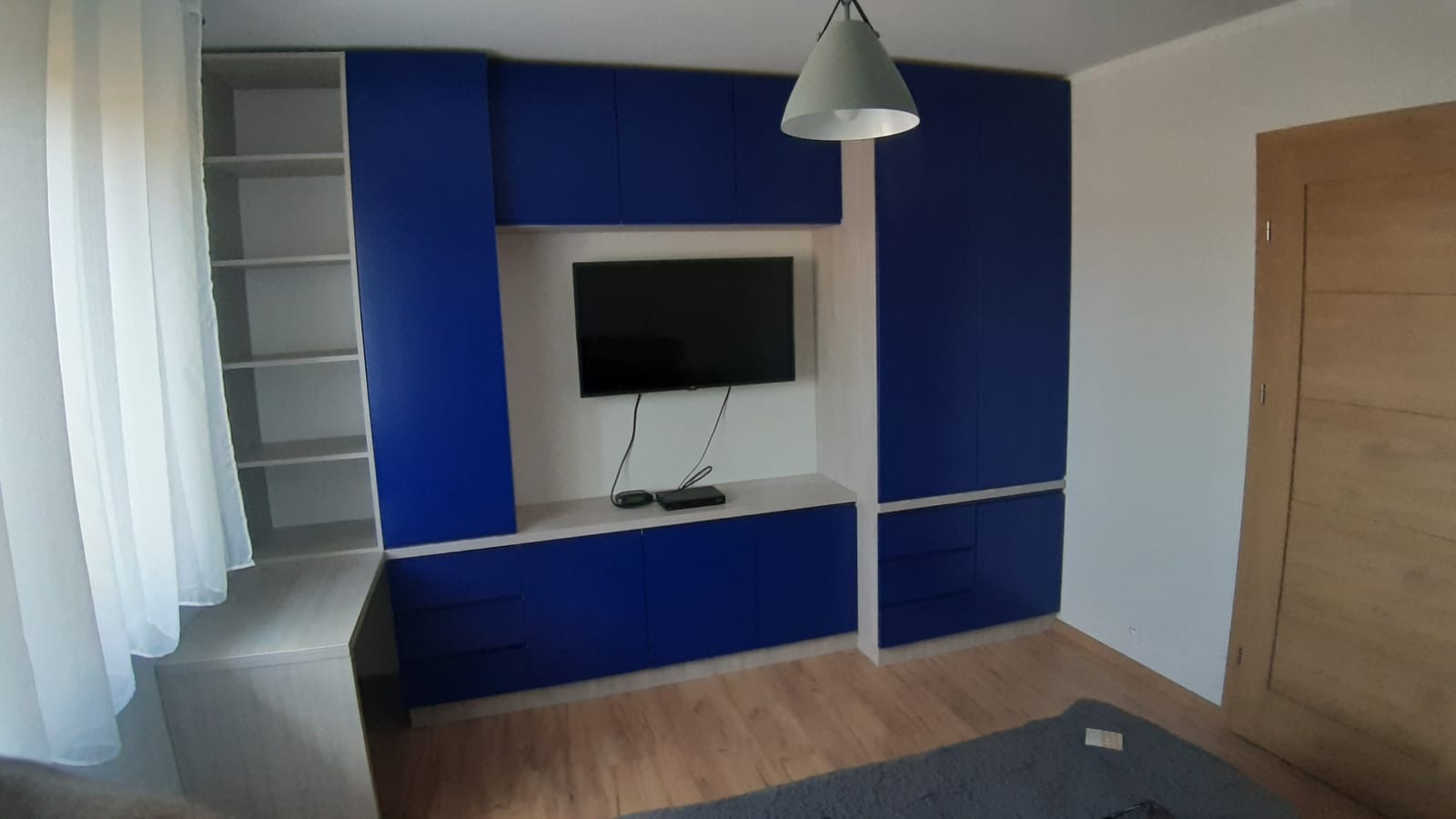 Niebieska zabudowa meblowa z szafkami, półkami i miejscem na telewizor w salonie w Słupsku, z widoczną lampą wiszącą i zasłoniętym oknem.