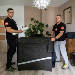 Dwóch pracowników firmy przeprowadzkowej w czarnych koszulkach z logo, zabezpiecza mebel czarną pikowaną osłoną i folią stretch w jasnym pokoju z roślinami.