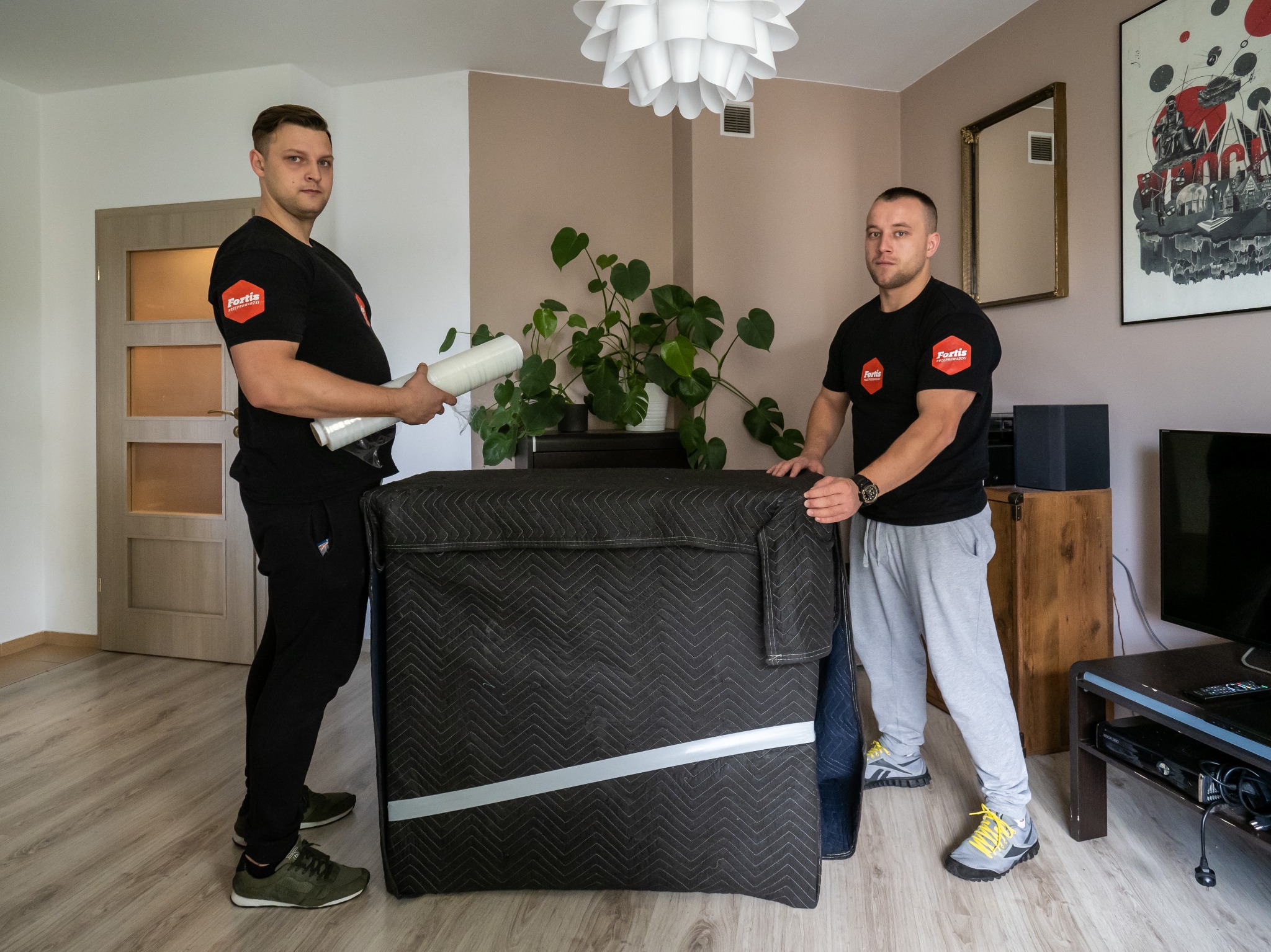 Dwóch pracowników firmy przeprowadzkowej w czarnych koszulkach z logo, zabezpiecza mebel czarną pikowaną osłoną i folią stretch w jasnym pokoju z roślinami.
