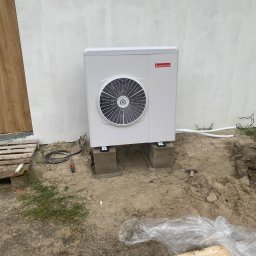Bartosz Racz Usługi hydrauliczne i gazowe - Biała pompa ciepła Ariston zamontowana na betonowych blokach przed jasną ścianą budynku. Widoczne przewody i narzędzia instalacyjne.