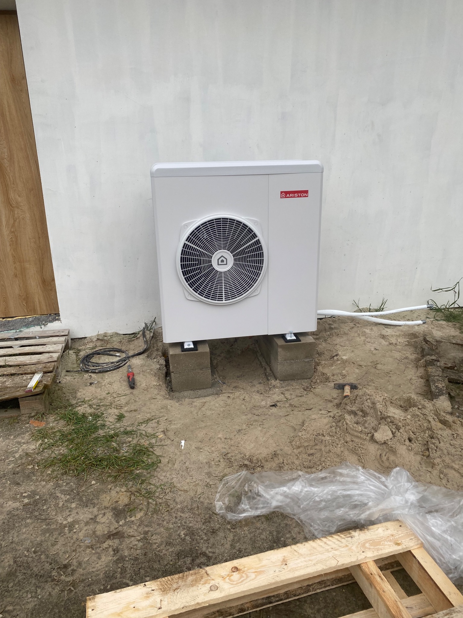 Biała pompa ciepła Ariston zamontowana na betonowych blokach przed jasną ścianą budynku. Widoczne przewody i narzędzia instalacyjne.