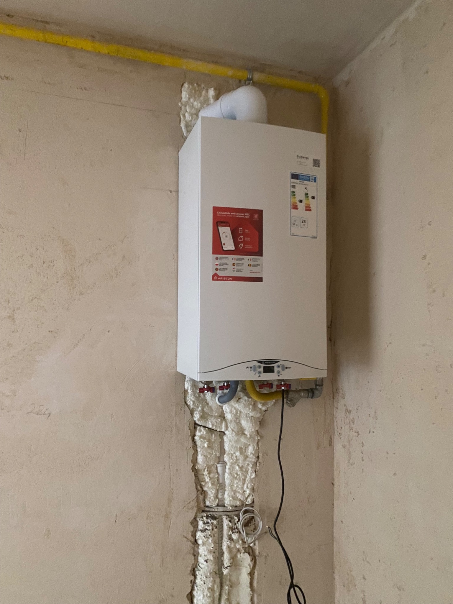 Biały, wiszący piec gazowy Ariston zamontowany na ścianie z widoczną instalacją i pianką montażową. Żółta rura gazowa biegnie wzdłuż sufitu. Widoczne przyłącza.