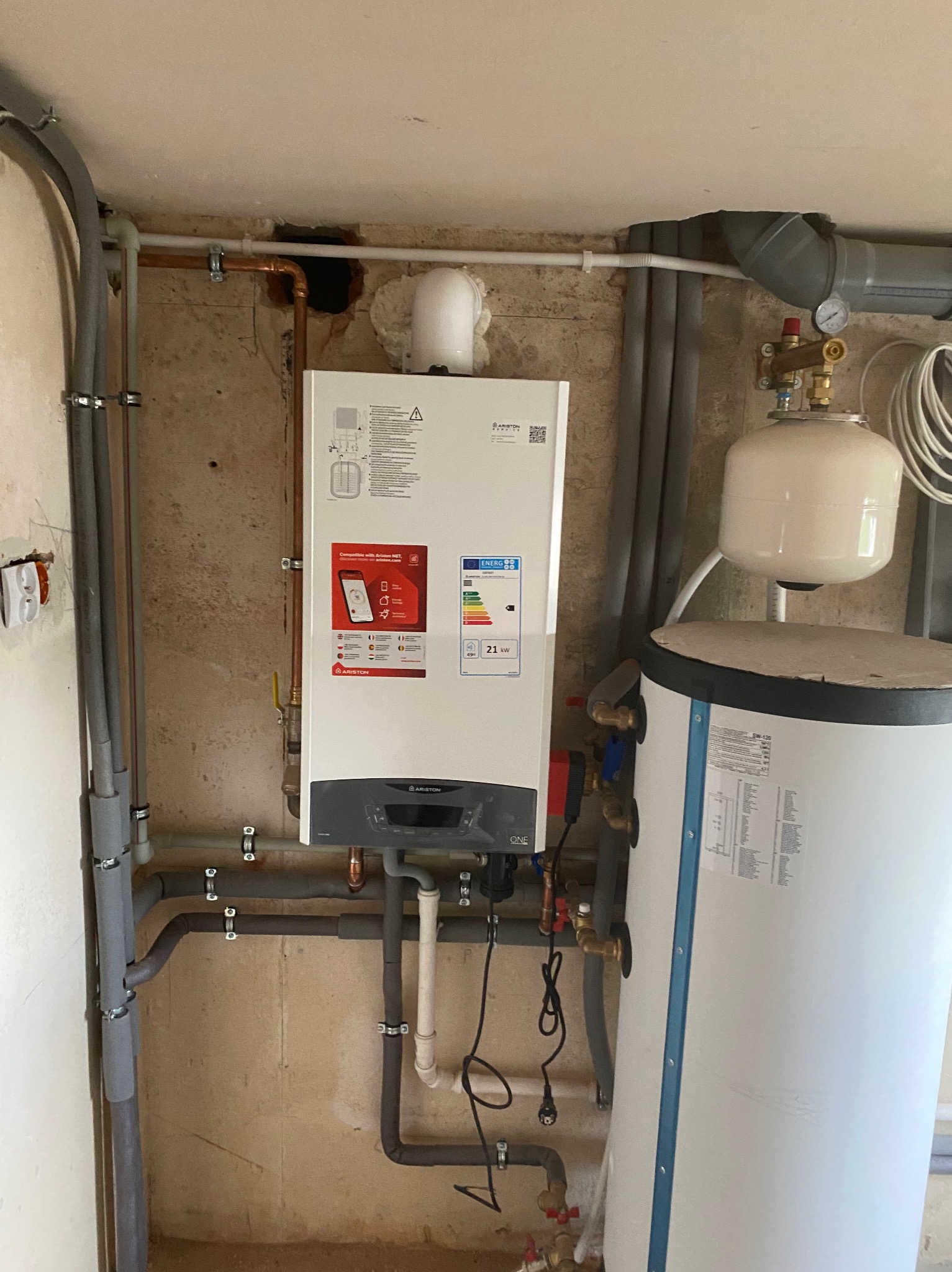Biały, wiszący kocioł gazowy Ariston z podłączonymi rurami miedzianymi i szarymi, oraz biały zbiornik na wodę, zamontowany na ścianie o surowym wykończeniu.
