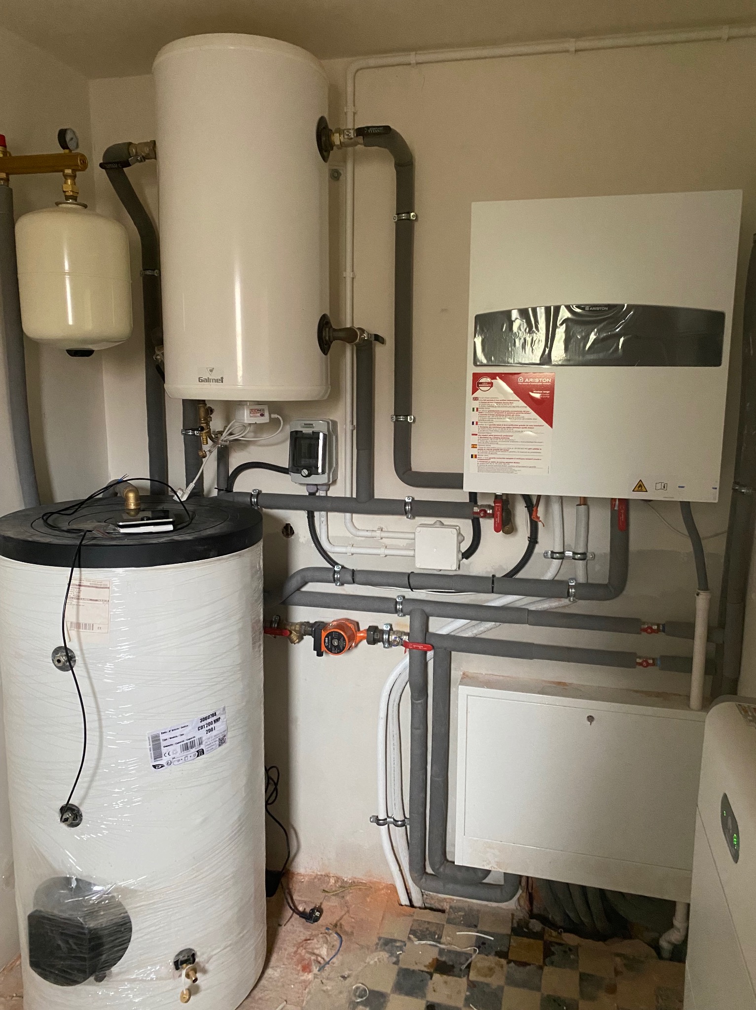 Biały, wiszący piec gazowy Ariston z szarymi rurami, zbiornikami i osprzętem na tle ściany. Widoczne elementy instalacji grzewczej w pomieszczeniu.