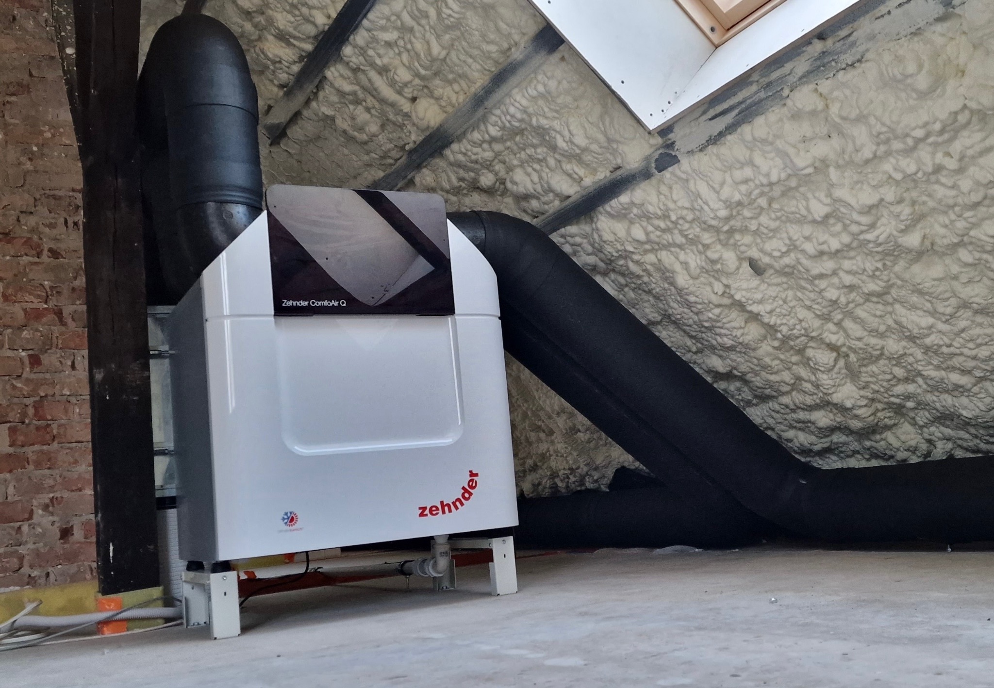 Biała jednostka rekuperacji Zehnder ComfoAir Q z czarnymi rurami wentylacyjnymi w pomieszczeniu z izolacją piankową na poddaszu. Widoczny fragment ceglanej ściany.